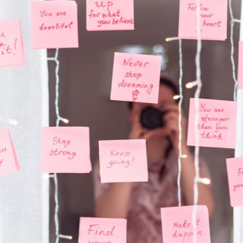 a mirror and a woman looking into it with pink sticky notes filled with words of affirmation
