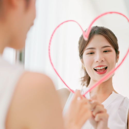 a woman smiling in the mirror and she has drawn a pink love heart around her face suggesting she has high self-worth